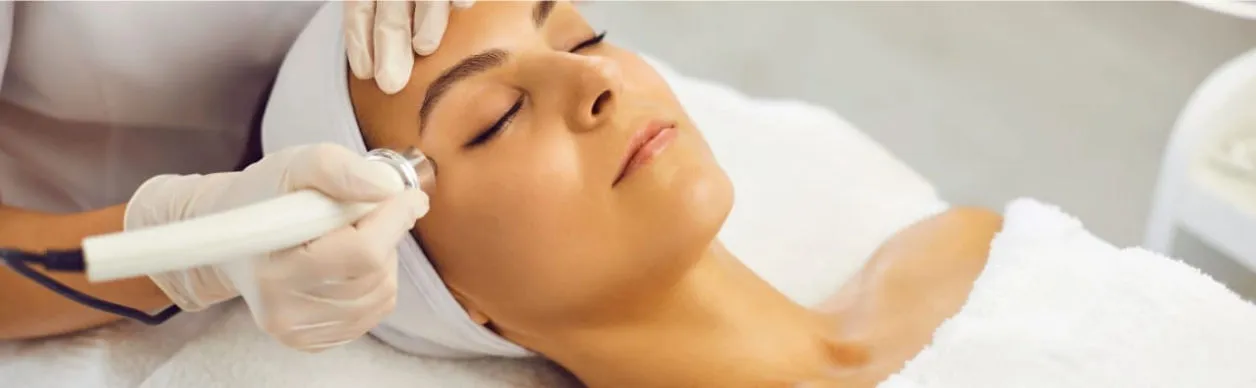Profissional aplicando aparelho de radiofrequência no rosto de uma cliente deitada em maca, em atendimento de estética facial